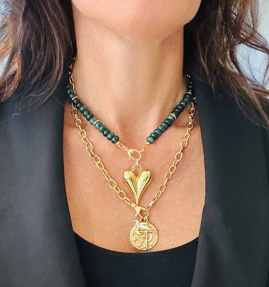 Gold necklace with heart pendant and green beaded chain worn by a person against a black background